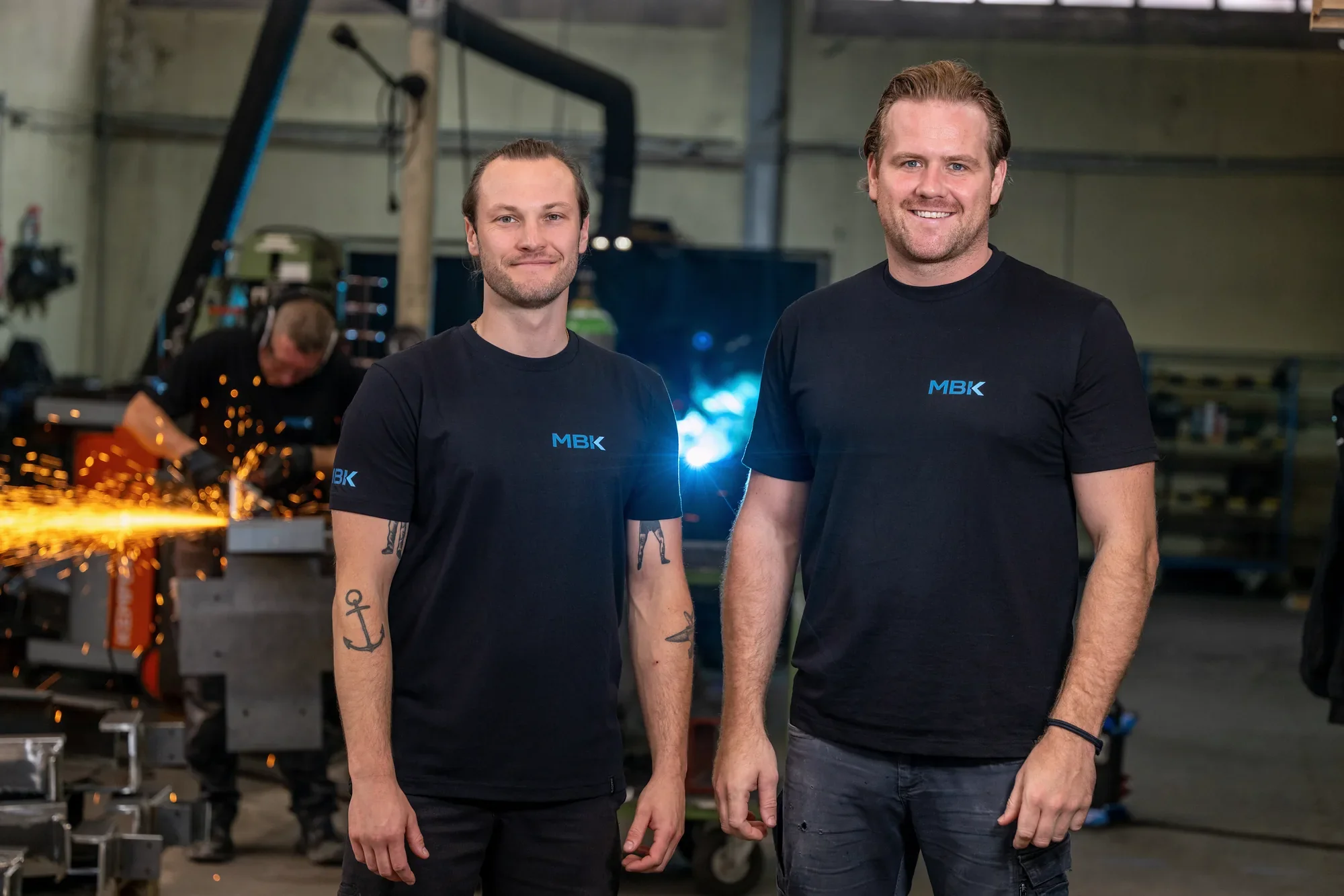 Die Geschäftsführer der MBK Metallbau Köln GmbH, dahinter ein Schweißer bei der Arbeit